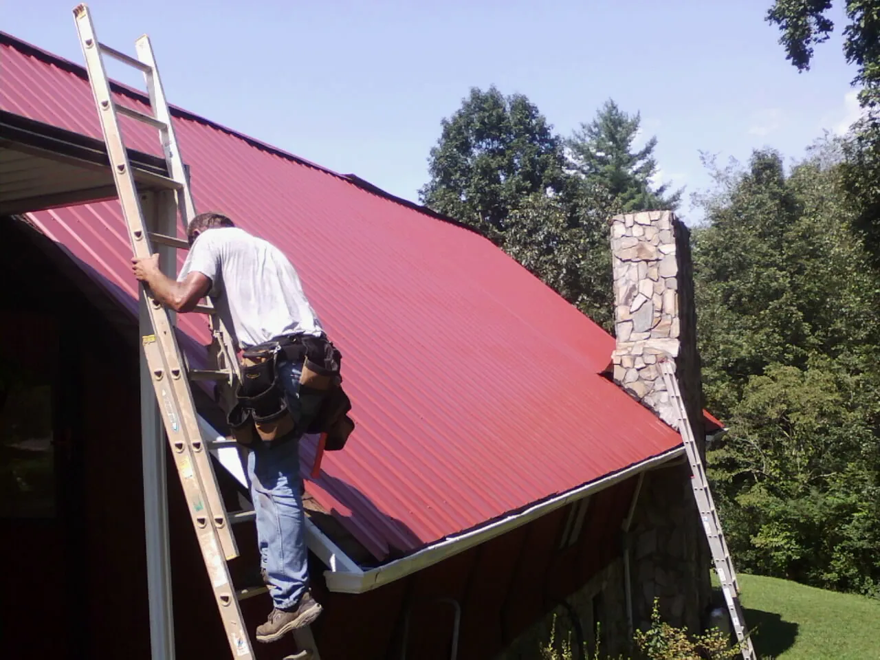 Roofer on a ladder beside a red metal roof and stone chimney — Soffit & Fascia Repair by Roofing Brothers in Roselle Park